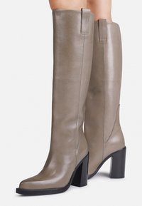 Bottes en cuir taupe jusqu'aux genoux avec des talons carré noirs portées sur des jambes, présentées sur un fond blanc.