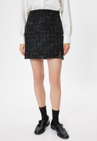 Sort tweed nederdel med hvide spidser, i en lige form med sideslids. Kombinationen vises med sorte loafers og sokker.