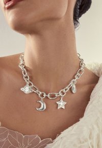 Silver chain necklace with planet, crescent moon, star, and teardrop charms worn on a woman's neck over a light lacy top and fluffy white garment.