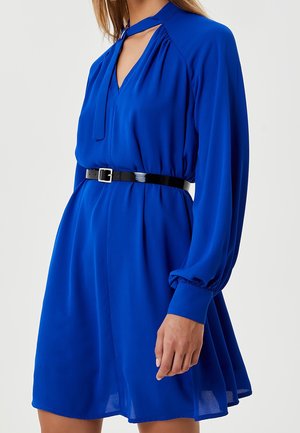Femme portant une robe bleu cobalt à manches longues avec un décolleté ajouré et une ceinture en cuir verni noir à la taille.