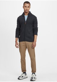 Chunky mørkeblå strikket cardigan med høj krave, struktureret overdel og ribbede ærmer, kombineret med brune bukser og sneakers.