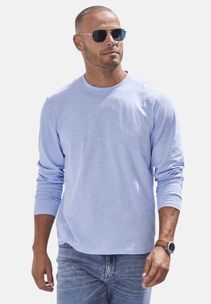 Lichtblauw overhemd met lange mouwen en een ronde hals, zachte textuur, gecombineerd met denim jeans en ronde zonnebril, wat een casual uitstraling geeft.