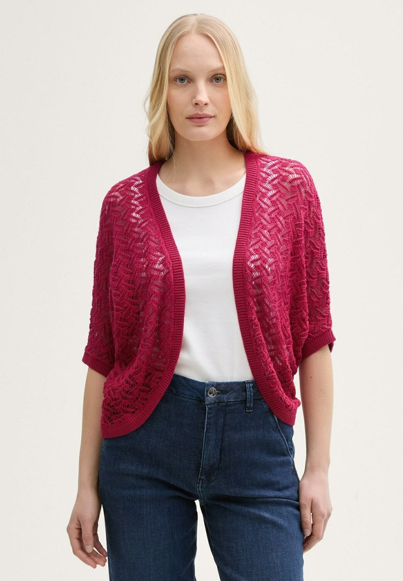 Cardigan rouge en tricot avec un design ouvert et des manches courtes, présentant un motif texturé. Porté sur un haut blanc et associé à un jean bleu.