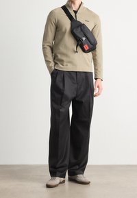 Man som bär en beige zip-up-tröja, löst sittande svarta byxor, grå sneakers och en svart axelväska med röd etikett, stående på ett ljust golv.