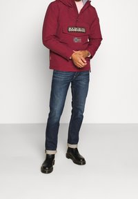 Giacca pullover rossa con tasca frontale, decorata con una patch grafica e la bandiera della Norvegia. Indossata con jeans blu e stivali neri con cuciture gialle.