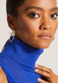 Rollkragenpullover aus leuchtend blauem Rippstrickmaterial, mit einem hohen Kragen und glatter Textur, getragen mit einem goldenen Creolen-Ohrstecker.