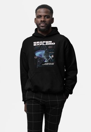 Uomo che indossa una felpa nera con cappuccio con grafica di un'astronave e la scritta "Semper Exploro" e pantaloni neri con motivo a griglia bianca.