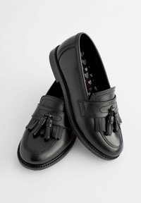 Paire de mocassins à pampilles en cuir noir avec détails de franges et doublure intérieure à motif de cœurs sur fond blanc.