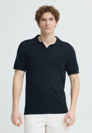 Ung mann med krøllete hår iført en svart kortermet polo og hvite bukser, stående mot en ensfarget lys grå bakgrunn.