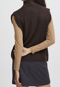 Pull sans manches à col roulé côtelé marron superposé sur une longue chemise beige, associé à une jupe gris foncé. Texture douce, coupe ajustée.
