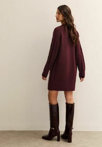 Vestito in maglia bordeaux con dettagli a treccia e orlo a costine, abbinato a stivali lucidi marroni con tacco alti fino al ginocchio.