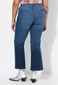 Jeans de talle alto de color azul oscuro, acampanados, con cinco bolsillos y costuras visibles, combinados con botas de tobillo plateadas brillantes. Tela de mezclilla texturizada.