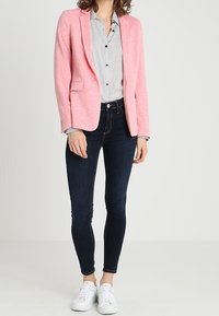 Blazer rosa chiaro con colletto a pistola, camicia a righe grigie, jeans skinny scuri e sneakers bianche. Tessuto morbido con vestibilità su misura.