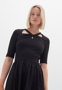 Schwarzes Kleid mit einem enganliegenden, gerippten Oberteil und kurzen Ärmeln, das über ausgeschnittene Details am Ausschnitt und einen fließenden, strukturierten Rock verfügt.