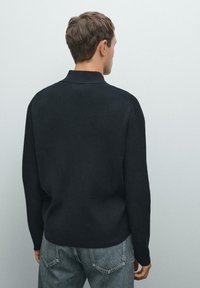 Uomo che indossa un maglione nero a coste e jeans blu, in piedi con le spalle alla fotocamera su uno sfondo grigio chiaro uniforme.