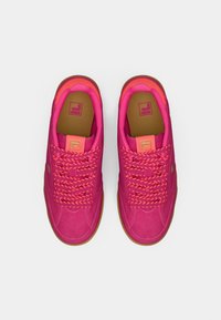 Rosa sneakers med et teksturert stofføvre, kontrasterende oransje snører og gummigummisåle. Logoen på tungen trykt i lys rosa.