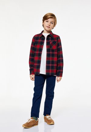 Camisa de franela a cuadros roja y azul marino con botones en el frente, combinada con una camiseta blanca, jeans de mezclilla azul y zapatillas marrones.