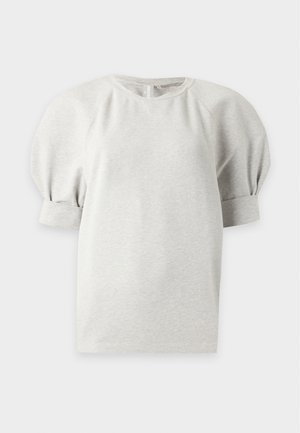 Femeie purtând un top gri deschis cu mâneci bufante, fustă mini neagră, sandale negre cu toc, ținând o geantă neagră din piele pe un fundal alb.