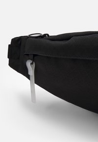 Svart midjeväska med en texturerad yta, prydd med en vit dragkedja och en cirkulär metallaccent. Kompakt och strömlinjeformad design.