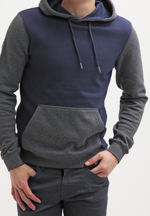 Persoon in een marineblauwe en grijze hoodie met een voorkantzak en koordjes, met de handen aan de taille, staand tegen een effen achtergrond.