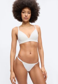 Γυναίκα με κατσαρά μαλλιά που φοράει λευκό σουτιέν και αντίστοιχο εσώρουχο, στέκεται μπροστά σε απλό φωτεινό φόντο, κοιτώντας προς τα εμπρός.