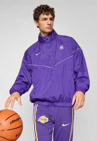 Lila Nike dzseki magas gallérral és cipzárral, fehér részletekkel. Sötétlila nadrággal párosítva, amelyen a "Los Angeles Lakers" logó látható.