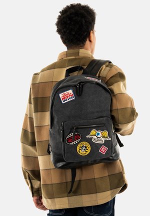 Sac à dos en tissu gris avec plusieurs écussons, doté de fermetures éclair, d'une poignée supérieure et de bretelles ajustables. Design texturé et décontracté avec des accents.