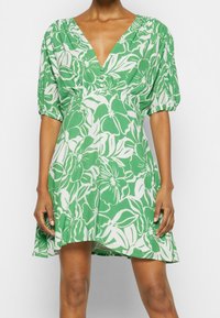 Žena v zelených a bílých květinových šatech s vycpanými rukávy a hlubokým výstřihem, stojící s uvolněnýma rukama podél těla.