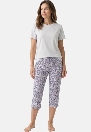 Vrouw in een lichtgrijs shirt met korte mouwen en paarse capribroek met bloemenprint en trekkoord, blootsvoets staand op een effen achtergrond.