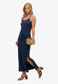 Granatowa maxi sukienka na ramiączkach, z bocznymi rozcięciami i gładką teksturą. Połączona z brązowymi sandałami na słupku i plecioną kopertówką.