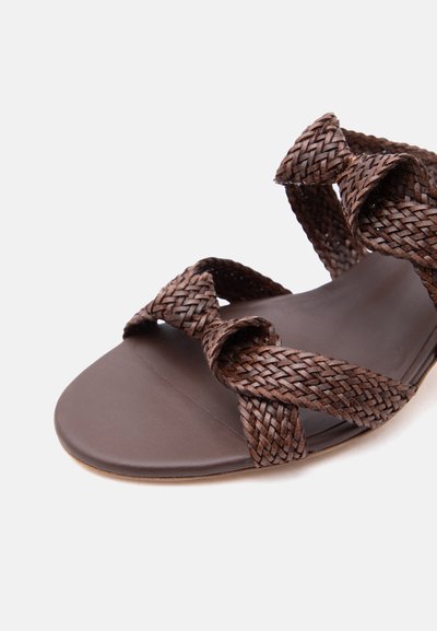 Καφέ σανδάλι με πλεγμένους δερμάτινους ιμάντες που διασταυρώνονται πάνω από το πόδι και μια επίπεδη, λεία σόλα σε λευκό φόντο.