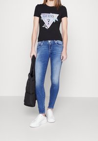 Czarna koszulka z grafiką trójkątnego logo w połączeniu z niebieskimi obcisłymi dżinsami. Obuwie sportowe w białym kolorze oraz czarna torba na ramię również są obecne.