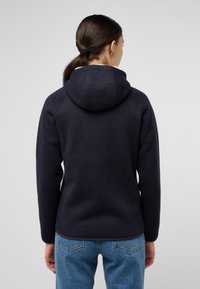 Navy gestrickter Kapuzenpullover mit strukturierter Oberfläche und einer breiten, weichen Kapuze. Kombiniert mit hellblauen Jeans, die ein lässiges Design und eine bequeme Passform zeigen.