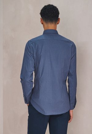 Uomo con capelli corti e ricci che indossa una camicia a quadri blu a maniche lunghe e pantaloni scuri, in piedi di spalle contro un muro semplice.