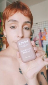 Una persona con i capelli rossi corti tiene un prodotto di trucco bronzer NYX Buttermelt vicino al viso in un ambiente interno.