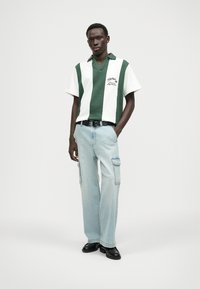 Zöld és fehér csíkos, rövid ujjú pólóing v-nyakkal, világoskék cargo nadrággal párosítva, amely oldalzsebekkel és fekete övvel rendelkezik.