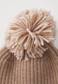 Un bonnet en tricot marron à côtes avec un grand pompon ouaté sur le dessus, sur fond blanc uni.