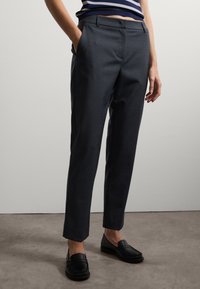 Dunkelgraue maßgeschneiderte Hose mit einem eleganten, figurbetonten Design. Verfügt über Seitentaschen und eine glatte Textur; kombiniert mit schwarzen Slippern.