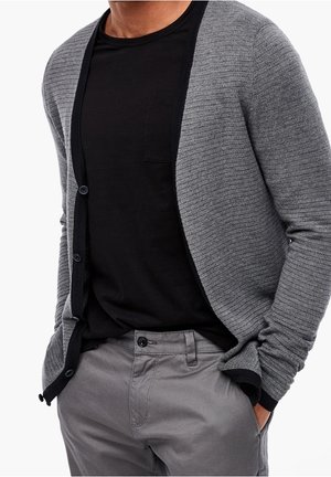 Grijze, gestructureerde cardigan met een contrasterende zwarte afwerking en knopen, gedragen over een zwart shirt, in combinatie met grijze broeken.
