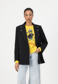 Blazer nero con dettagli in bottone argentato sopra un maglione giallo con design a cartone animato, abbinato a jeans a gamba larga di un blu chiaro.