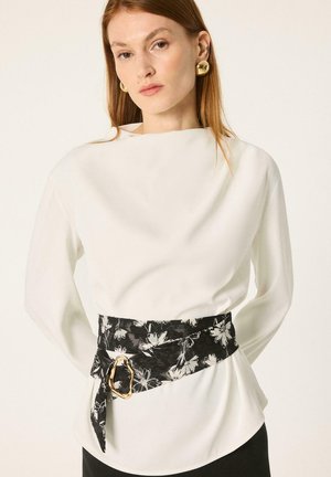 Mujer con cabello rojo liso lleva una blusa blanca de manga larga, grandes pendientes dorados y un cinturón ancho negro con flores y hebilla dorada irregular.