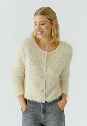 Lächelnde Frau mit kurzen blonden Haaren trägt eine flauschige cremefarbene Jacke mit Knöpfen und dunkelgraue Jeans, die Hände in den Taschen, vor einem schlichten hellen Hintergrund.
