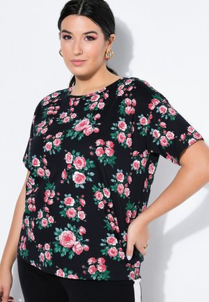 Vrouw met donker haar die gouden oorringen draagt en een zwarte top met korte mouwen met roze rozenprint, poserend met één hand op haar heup.