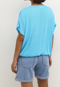 Personne portant un haut ample à manches courtes bleu clair et un short en jean retroussé, vue de dos sur un fond uni.