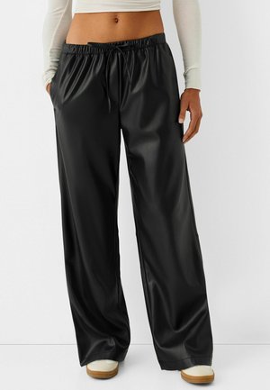 Femme portant un pantalon noir ample en faux cuir taille haute avec une ceinture élastique et un cordon de serrage, associé à des baskets blanches et un haut court à manches longues.