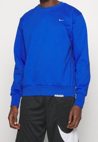 Personne portant un sweat-shirt Nike bleu royal et un short Nike noir avec un grand swoosh blanc, debout devant un fond uni.