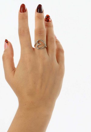 Silberner Ring mit offenem Herzmotiv, getragen an einer Hand mit mehrfarbigem Nagellack. Die Finger sind leicht gespreizt, um die Form des Rings zu zeigen.