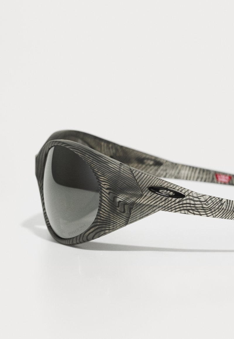 Occhiali da sole con montatura grigia a fantasia e lenti nere. La texture unica ricorda le venature del legno, con un logo Oakley sul lato.