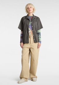 Short-sleeve, charcoal shirt with side pockets, button closure, over a multi-colored long-sleeve top; paired with beige, wide-leg trousers.