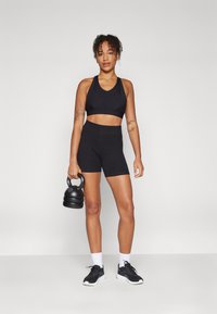 Svart sport-bh og matchende høyt liv-shorts, laget av elastisk stoff. Modellen holder en svart kettlebell, iført svarte joggesko og hvite sokker.